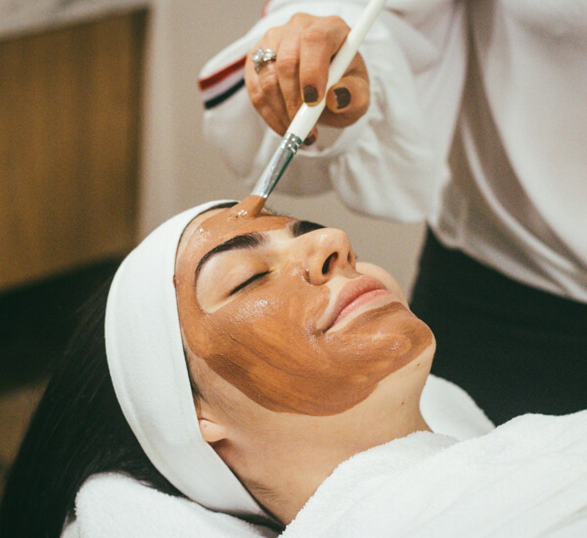 A therapist applying a facial mask to a client lying on a treatment bed